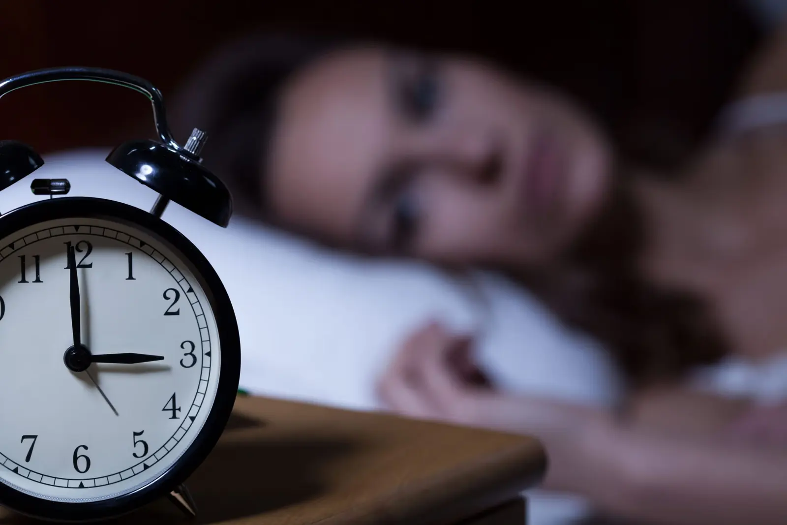 Woman lying awake in bed at night beside an alarm clock showing three o'clock.
