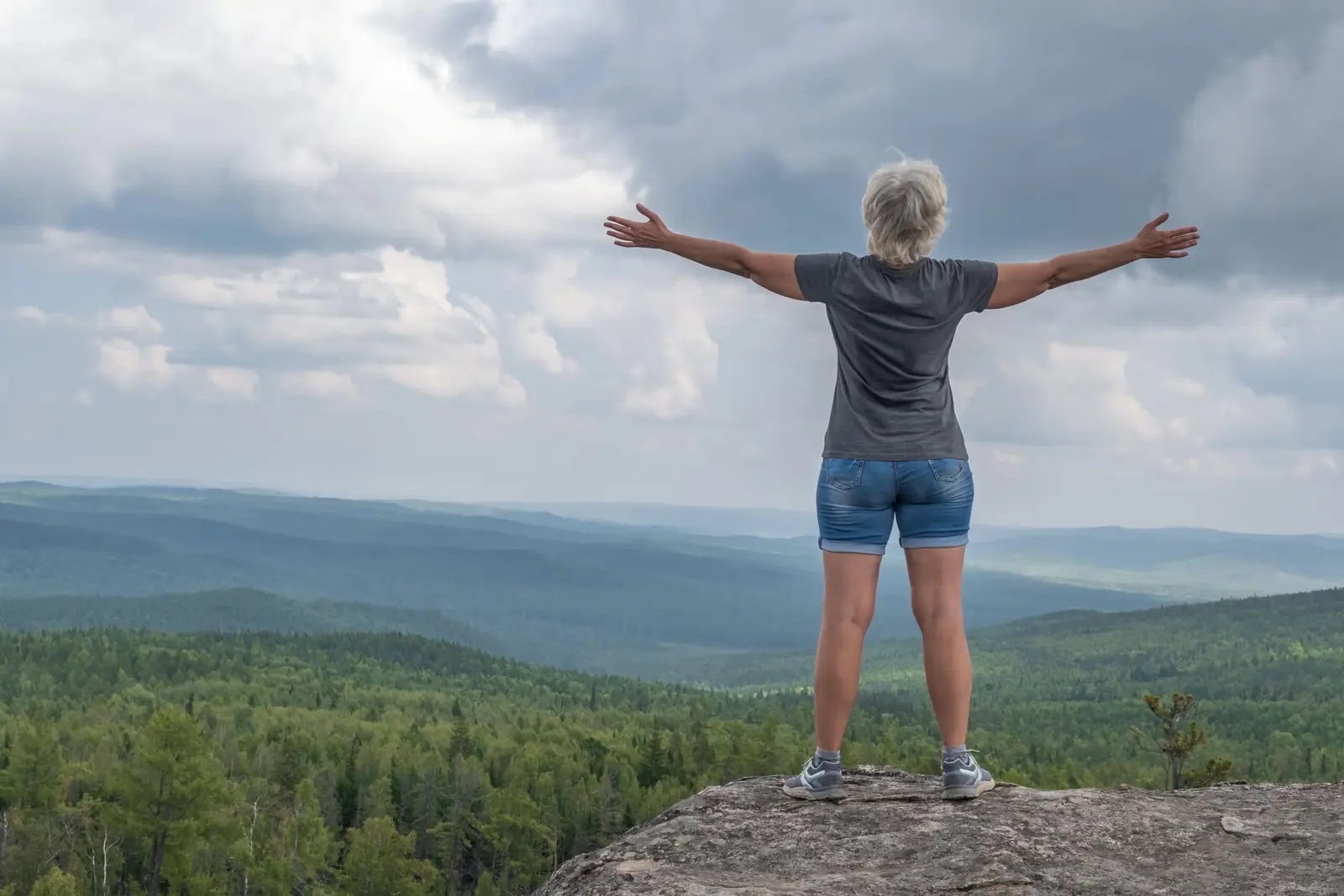Woman standing on a rocky hilltop with her arms outstretched above a forest valley.