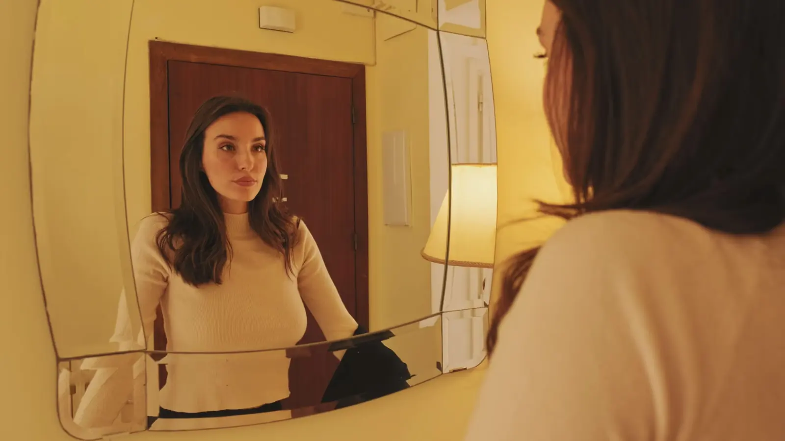 Woman with long dark hair looking at her reflection in a mirror.