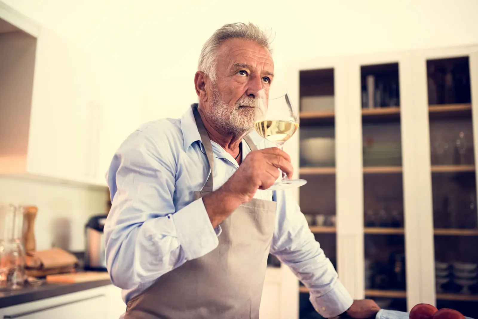 Older man in an apron drinking a glass of white wine in his kitchen.
