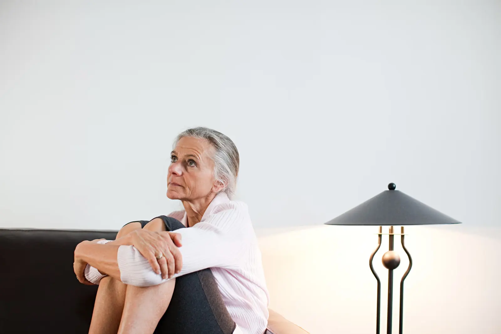 Older woman sitting on a sofa hugging her knees and looking up thoughtfully.