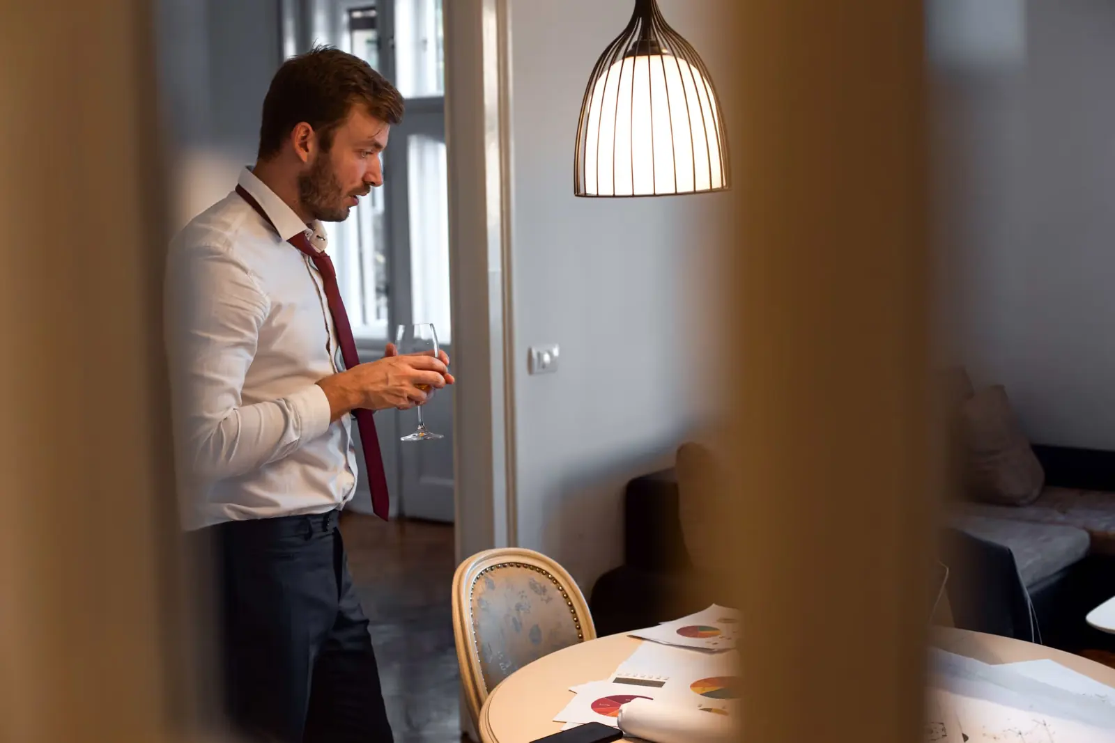 Man in a shirt and tie drinking a glass of wine at home, seen through a doorway.