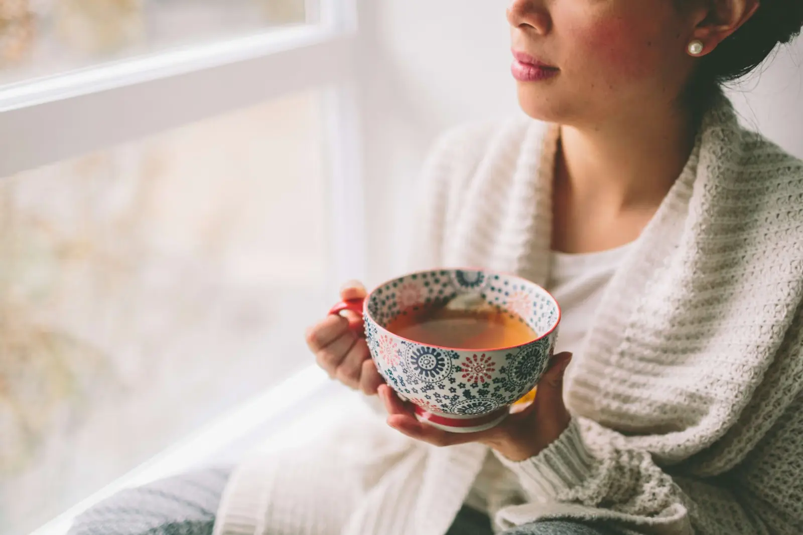 Woman in a cosy cardigan holding a cup of tea by a window.