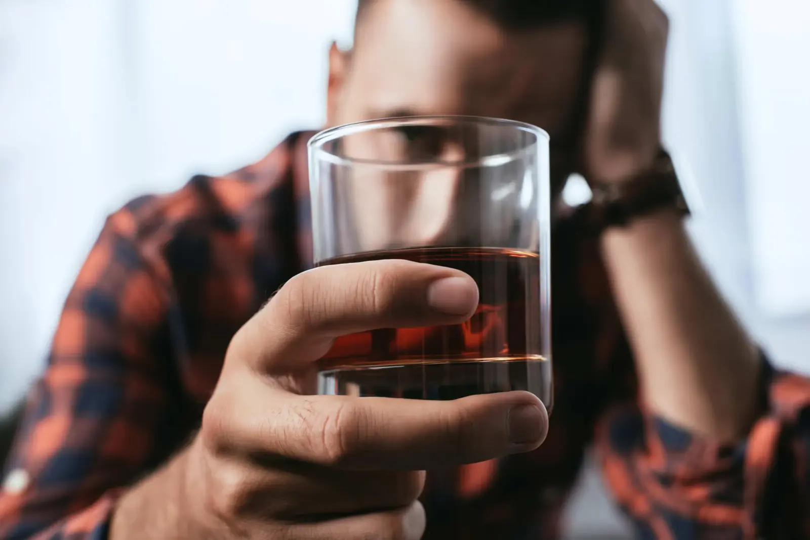 Man in a checked shirt holding up a glass of whisky with his head resting on his hand.