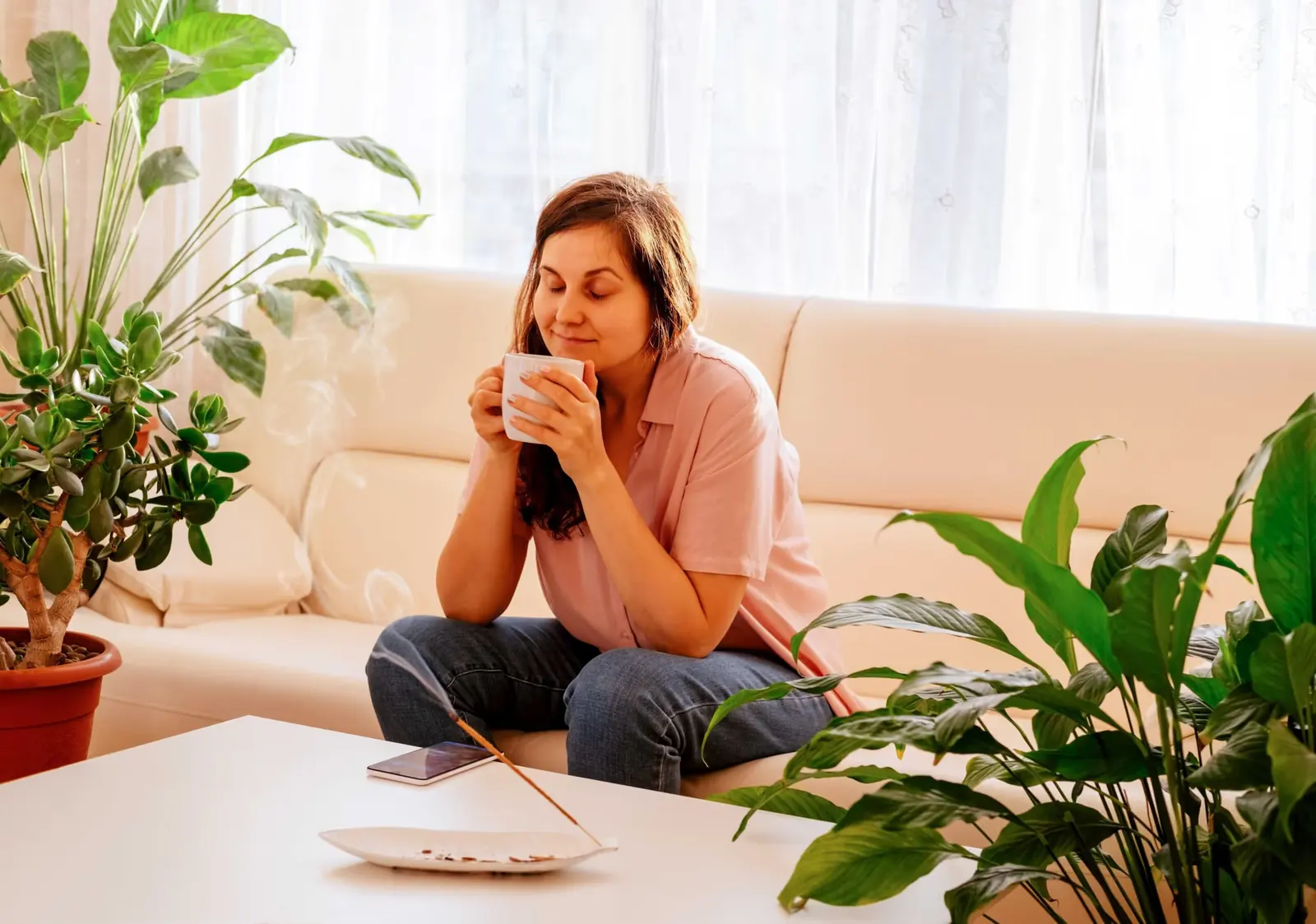 Woman drinking from a mug on a sofa surrounded by house plants.