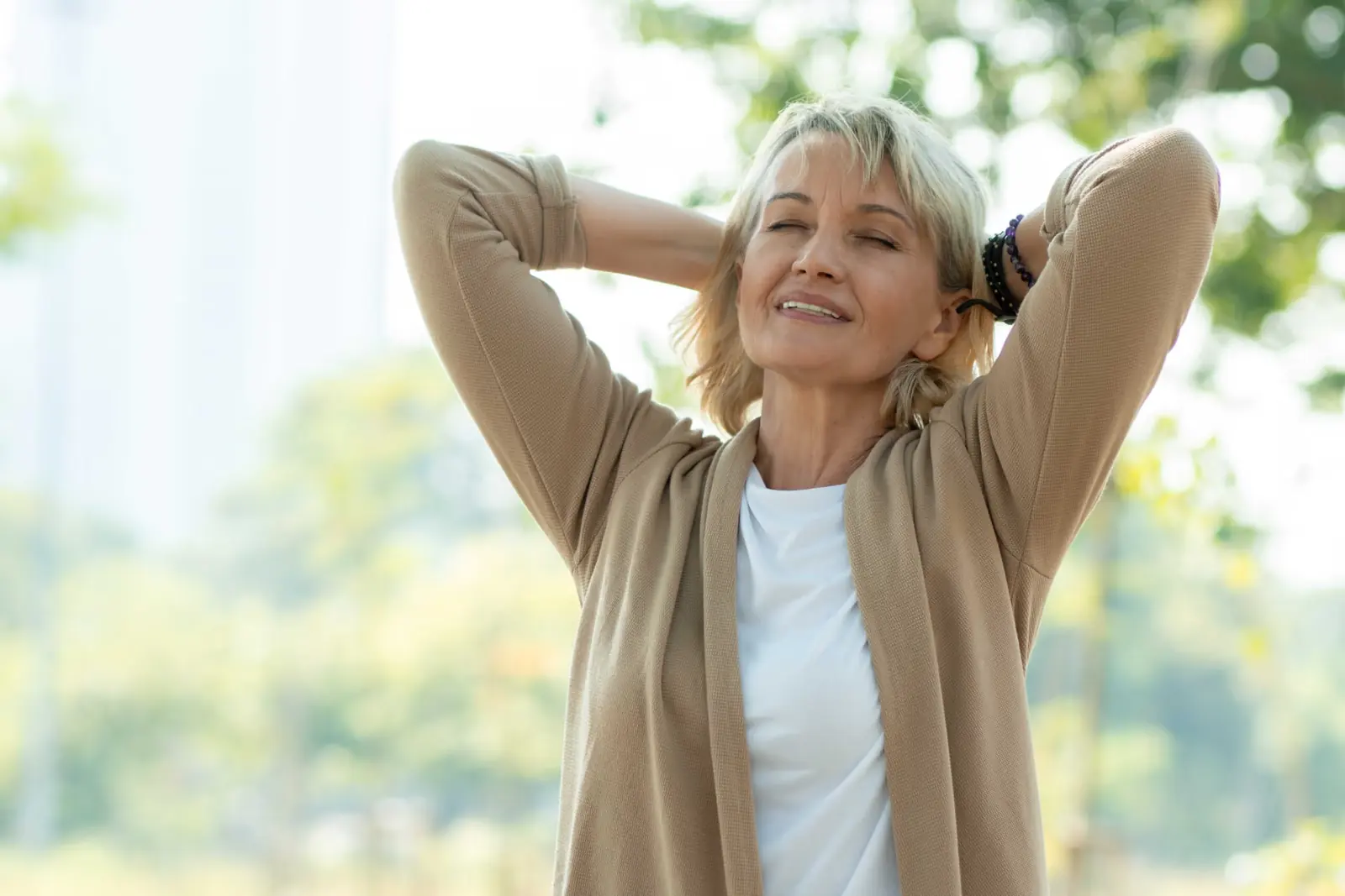 Relaxed older woman smiling outdoors with her hands behind her head.