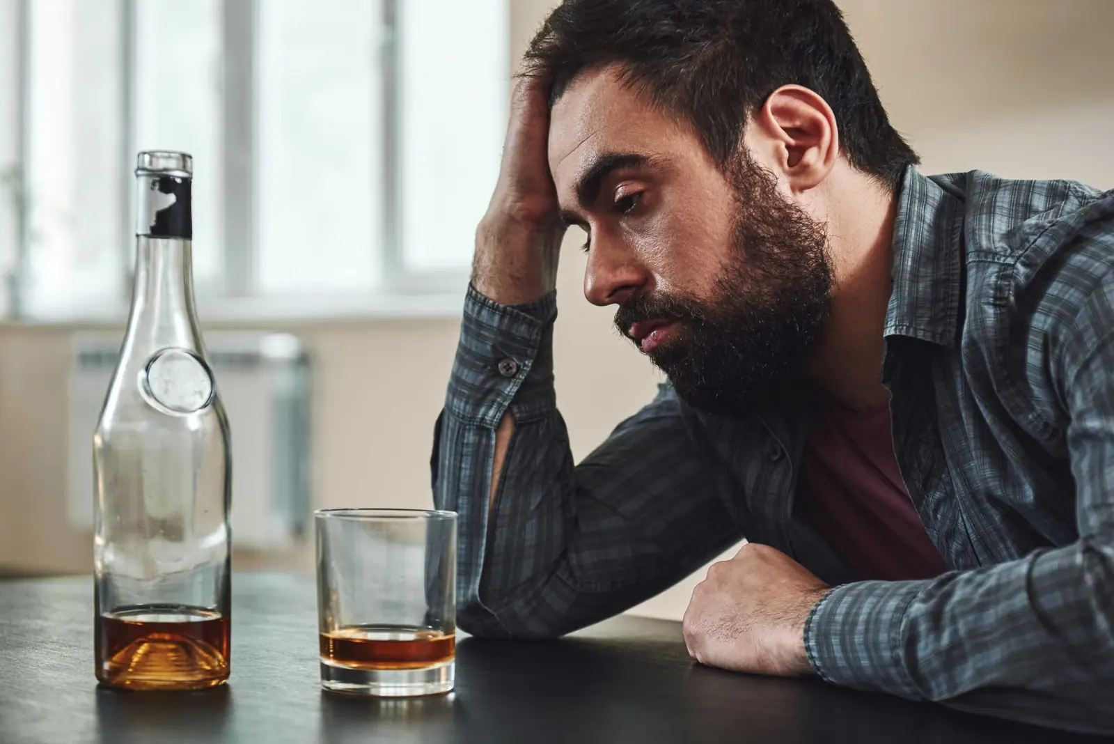 Stressed man holding his head at a table beside a bottle and a glass of whisky.