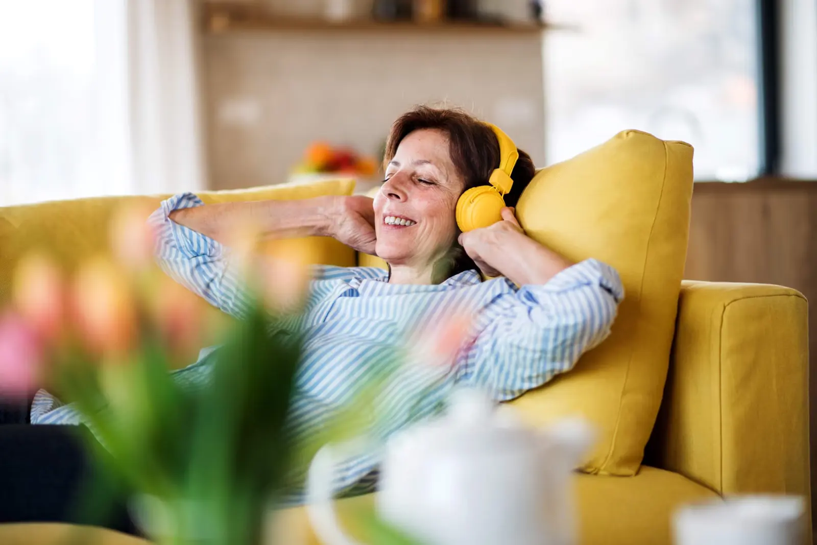 Smiling woman relaxing on a yellow sofa and listening to music on headphones.