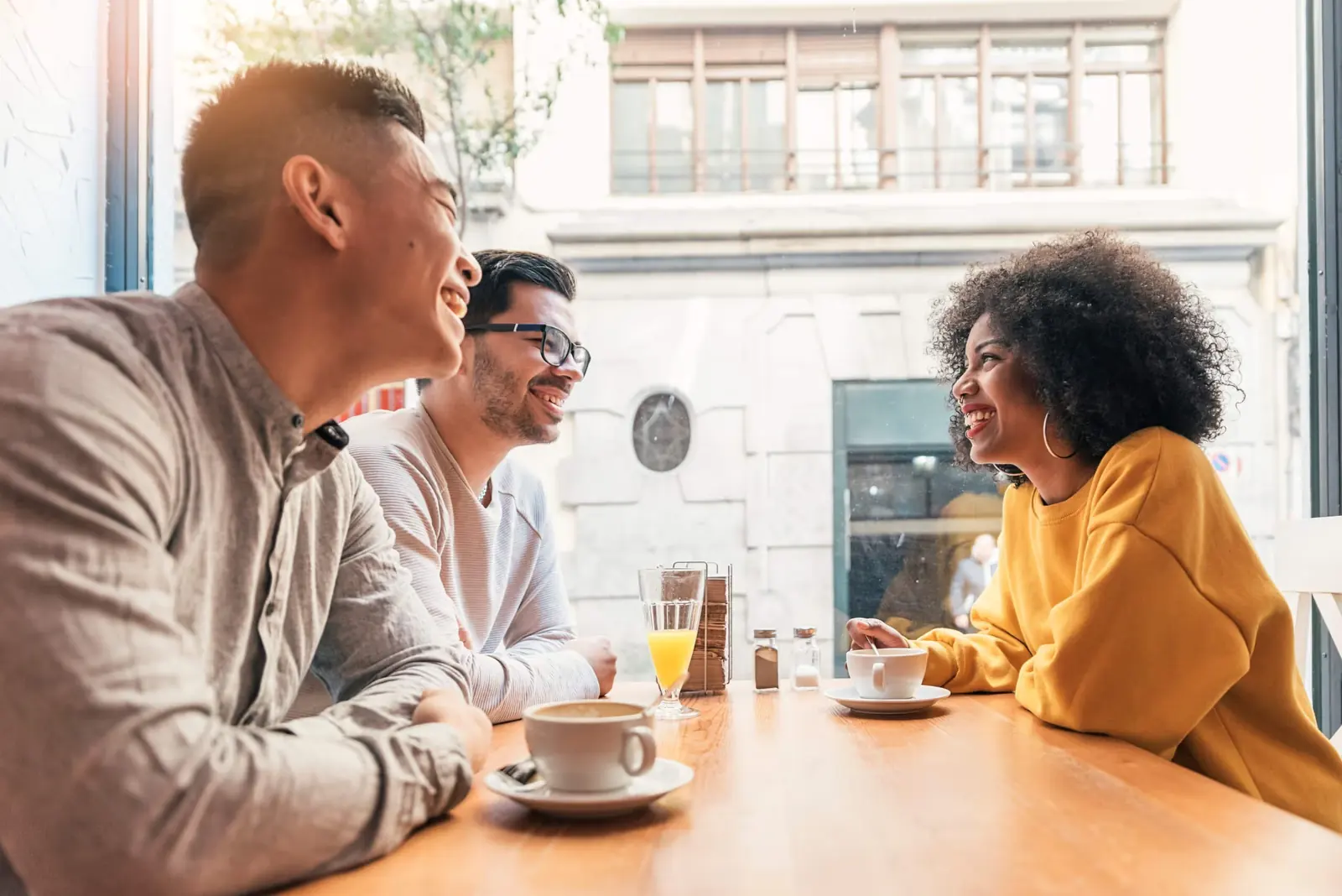 Friends laughing together over coffee and orange juice at a cafe table.