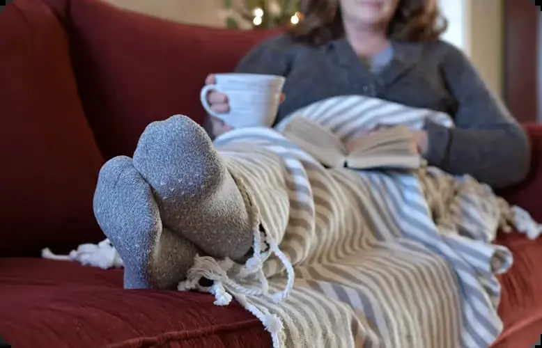 Woman curled up under a blanket on the sofa with a mug and a book.