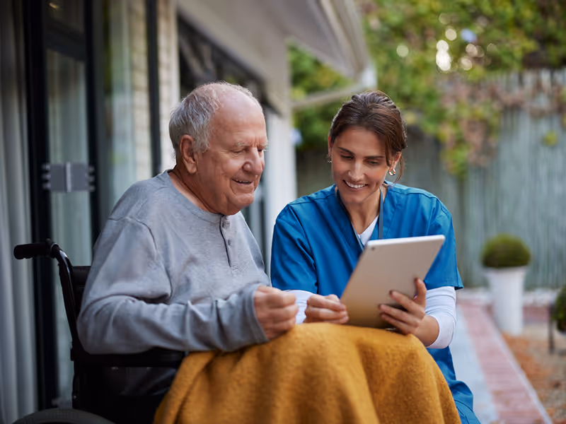 Therapeutin zeigt Patient im Rollstuhl draußen etwas auf einem Tablet.