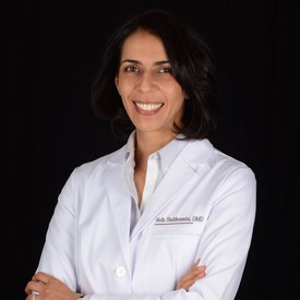 Smiling female doctor with dark hair wearing a white lab coat against a black background.