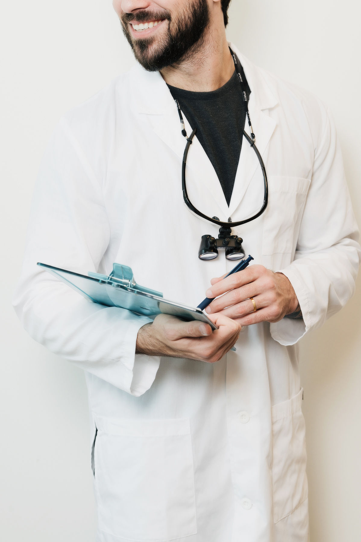 Smiling male doctor in white coat holding a clipboard and pen with magnifying loupes around his neck.