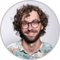 Smiling man with curly hair, beard, glasses, and a floral shirt against a light background.