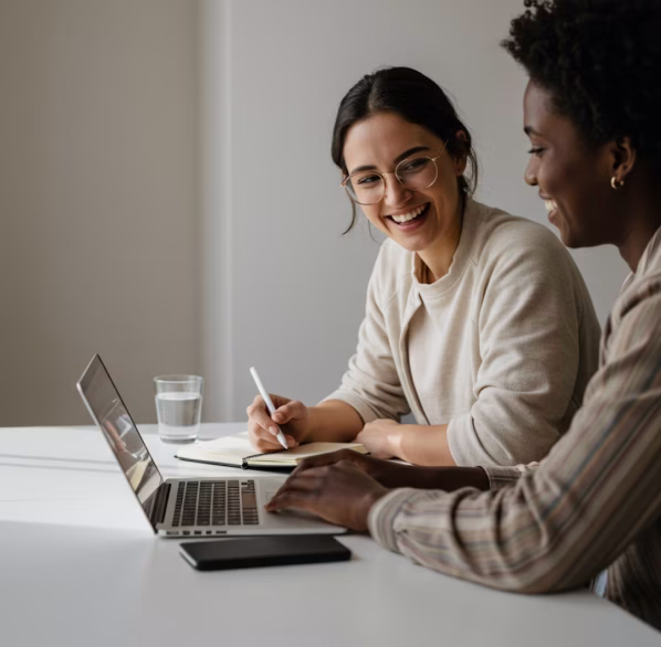 Two women smiling and collaborating while working on a laptop and taking notes at a white table.