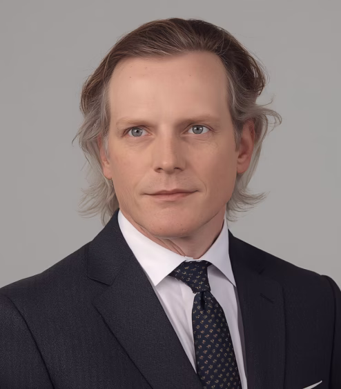 Portrait of a middle-aged man with light skin and grayish hair wearing a dark suit, white shirt, and patterned tie against a gray background.