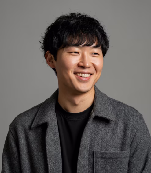 Smiling young Asian man with curly black hair wearing a dark gray coat and black shirt against a gray background.