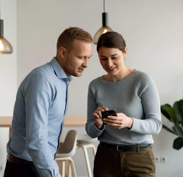 A man and a woman standing indoors, looking at a smartphone together and smiling.