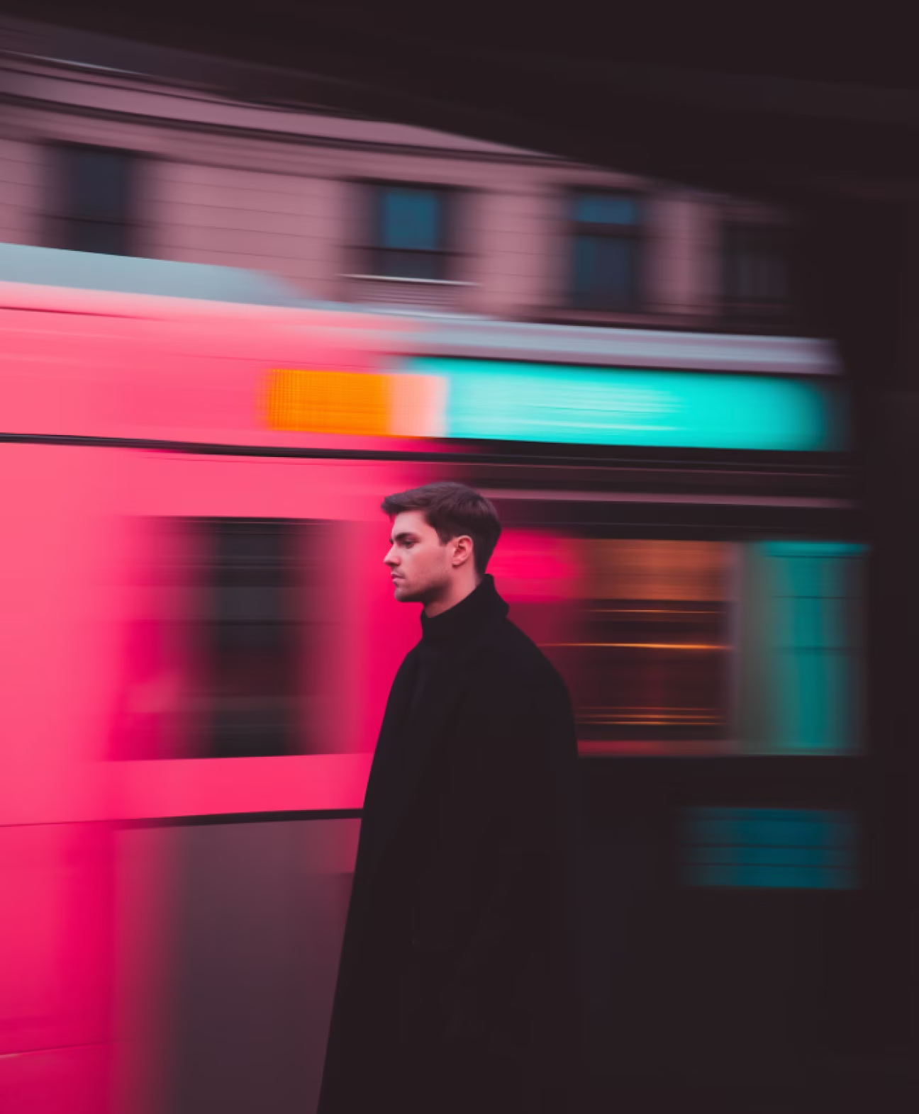 Profile of a young man in a black coat standing in front of a moving pink and teal tram.