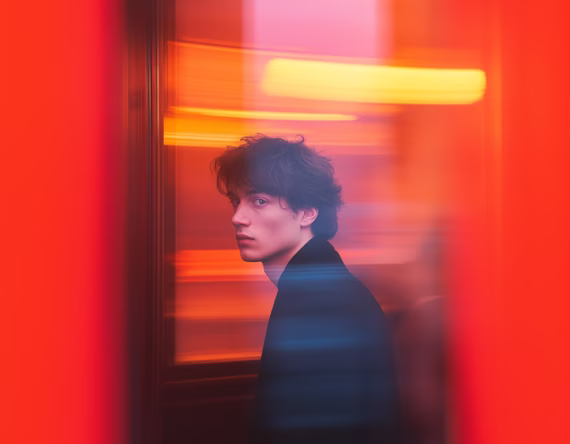 Young man with dark curly hair looking sideways, surrounded by red and orange blurred lights.