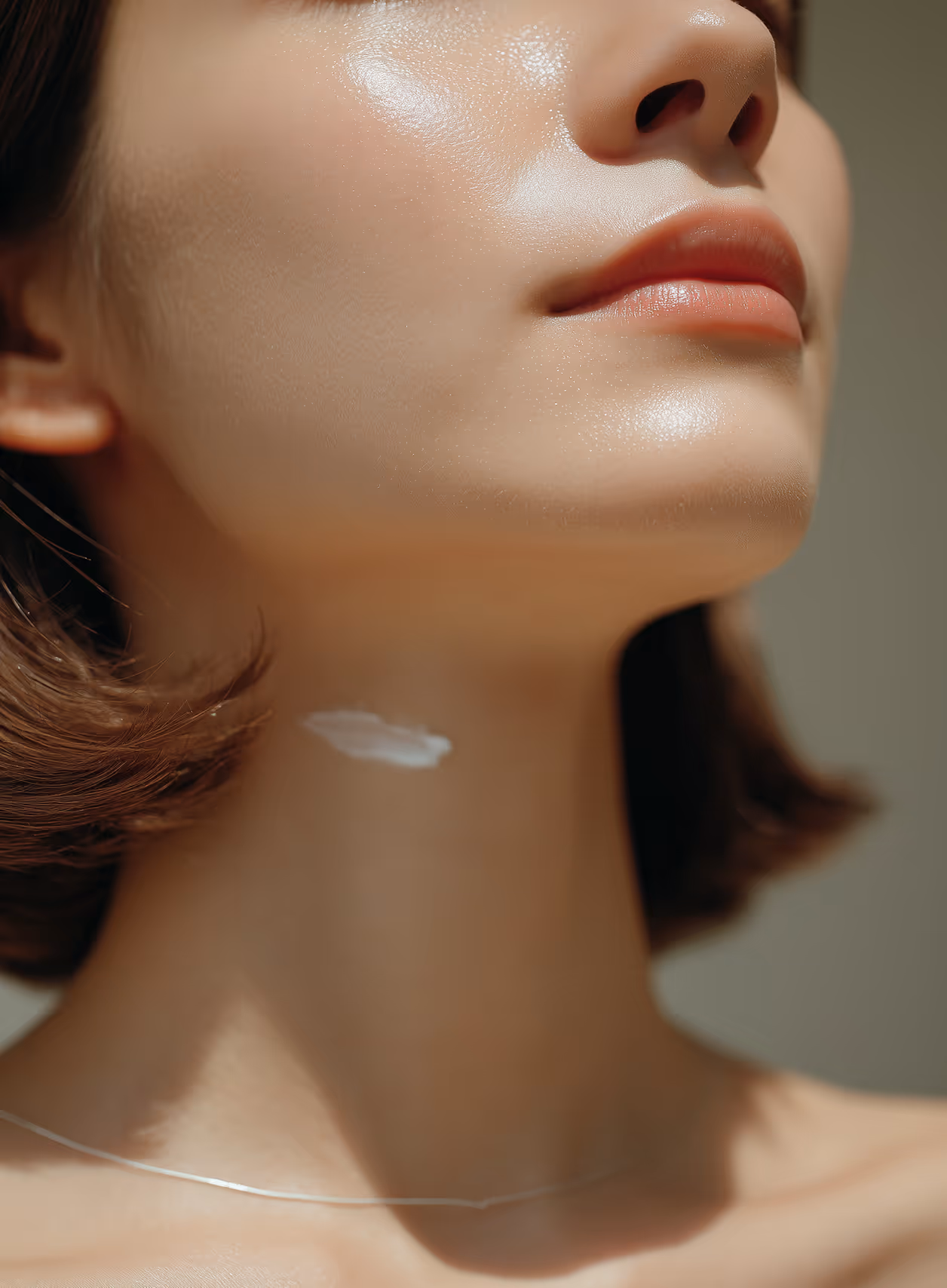 Close-up of a woman's lower face and neck with glowing skin and a small dab of white cream on her neck.
