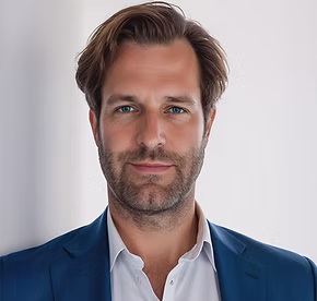 Portret van een man met een blauwe jas en wit overhemd, zittend voor een lichte achtergrond.