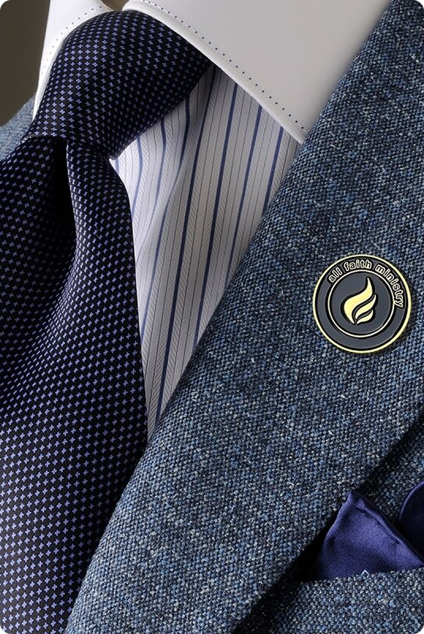 Close-up of a blue textured suit jacket with a round enamel pin, striped white and blue dress shirt, and dark blue patterned tie.
