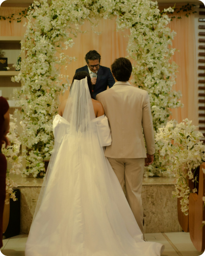 Bride and groom standing together facing an officiant under a large floral arch during a wedding ceremony.