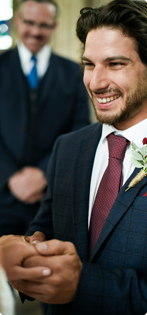 Smiling groom in a dark suit and red tie holding hands with a partner during a wedding ceremony.