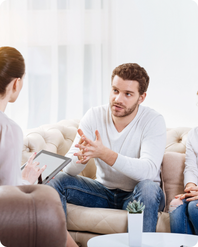 Man in white sweater discussing with a woman holding a tablet on a beige sofa in a bright room.