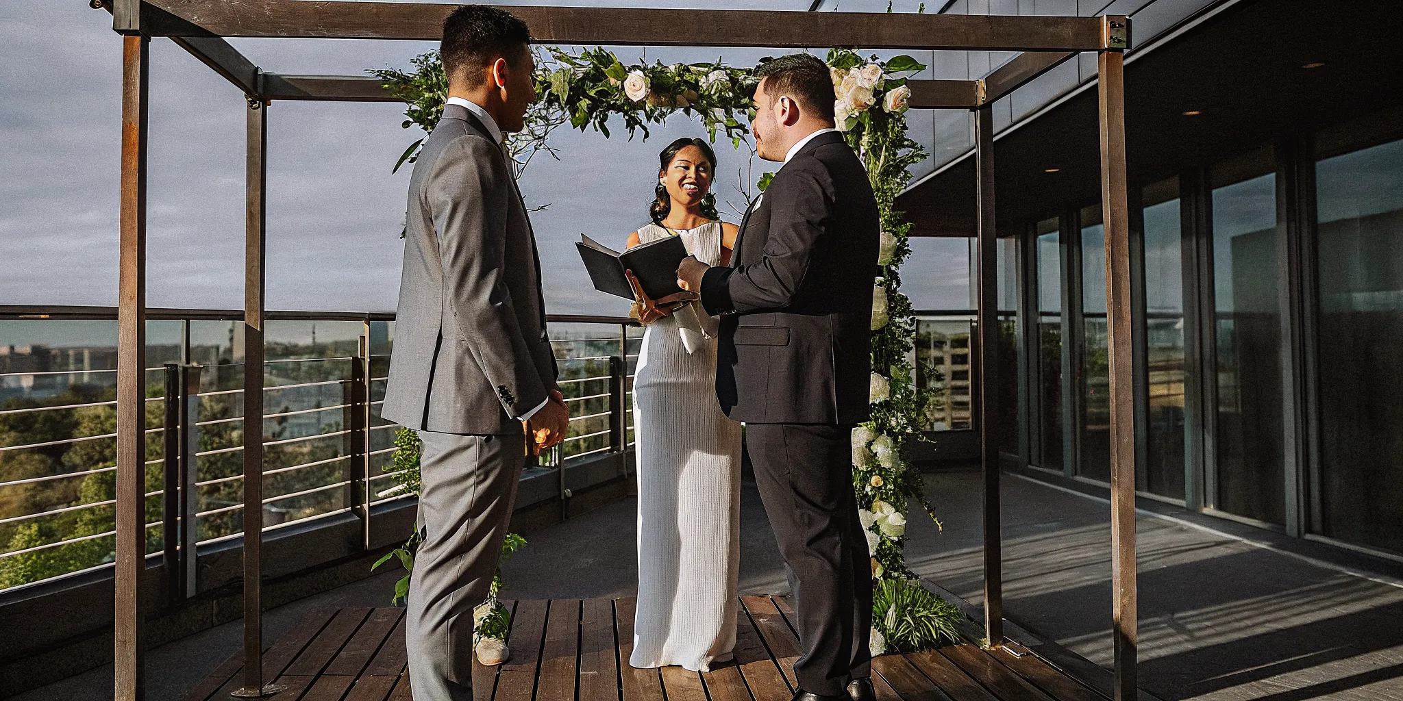 A good wedding officiant leads a rooftop ceremony for two grooms under a floral arch.