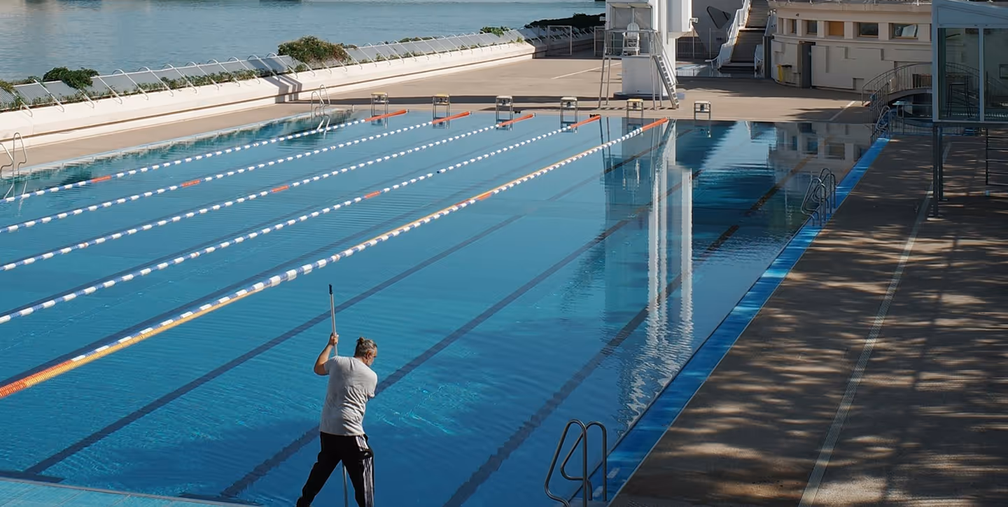 Person cleaning an empty outdoor swimming pool with a large pole during daylight.