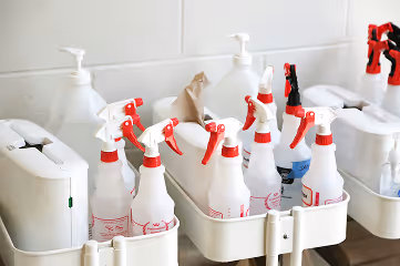 Several white spray bottles with red nozzles neatly organized in white holders on a metal cart against a white wall.