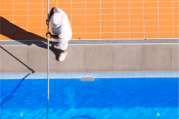 Overhead view of a person cleaning a pool with a long pole beside a blue swimming pool and orange tiled deck.