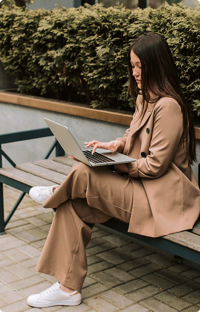 Woman in a brown suit sitting on a bench outdoors using a laptop.