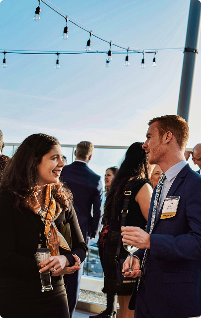 Two professionals, a woman and a man in a suit holding drinks, engaged in conversation at an outdoor networking event with string lights overhead.