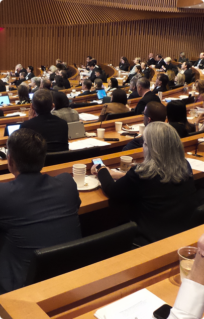 People attending a conference or meeting seated in a large auditorium with wooden paneling, using laptops and smartphones.