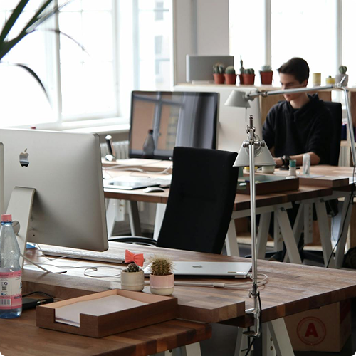 Modern office desk with iMac, laptop, small potted plants, and a person working at a computer in the background.
