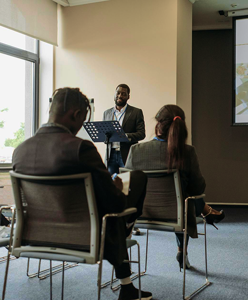 Man standing and speaking at a lectern in a conference room with two people seated facing him.