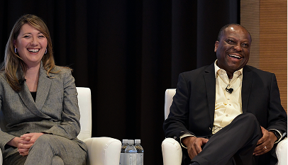 Two business professionals laughing while seated in white chairs during a panel discussion or interview.
