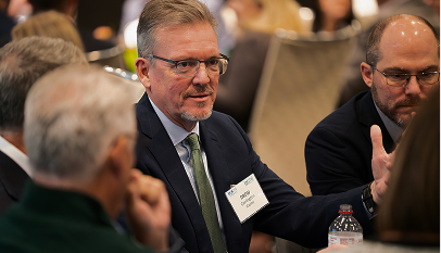 Two men in business attire engaged in conversation at a meeting table with blurred background attendees.