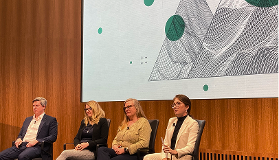 Four people sitting on chairs on a stage, in front of a wooden wall and a large screen displaying abstract geometric shapes.