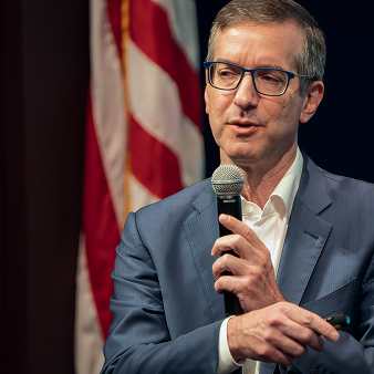 Man in a blue suit and glasses speaking into a microphone with an American flag in the background.