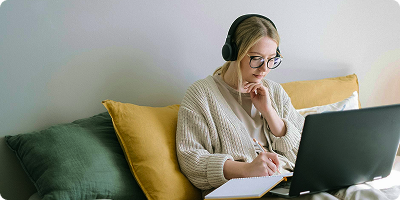 Young woman with glasses and headphones sitting on a couch, writing in a notebook while looking at a laptop.