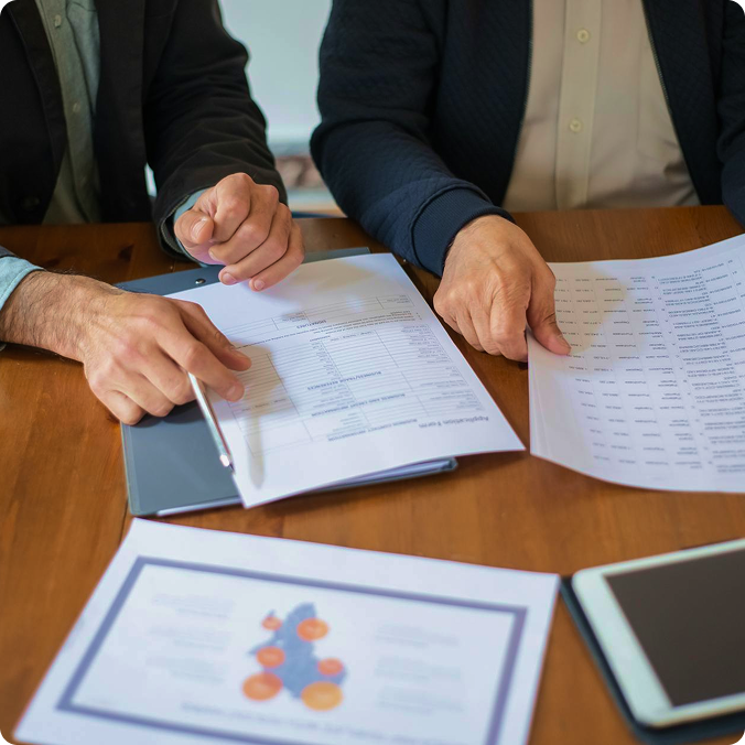 Two people reviewing and pointing at printed documents with charts and text on a wooden table.