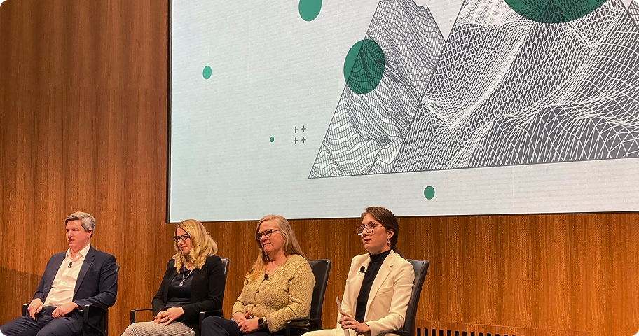 Four panelists seated on stage in front of a wooden wall and a screen displaying abstract geometric patterns.