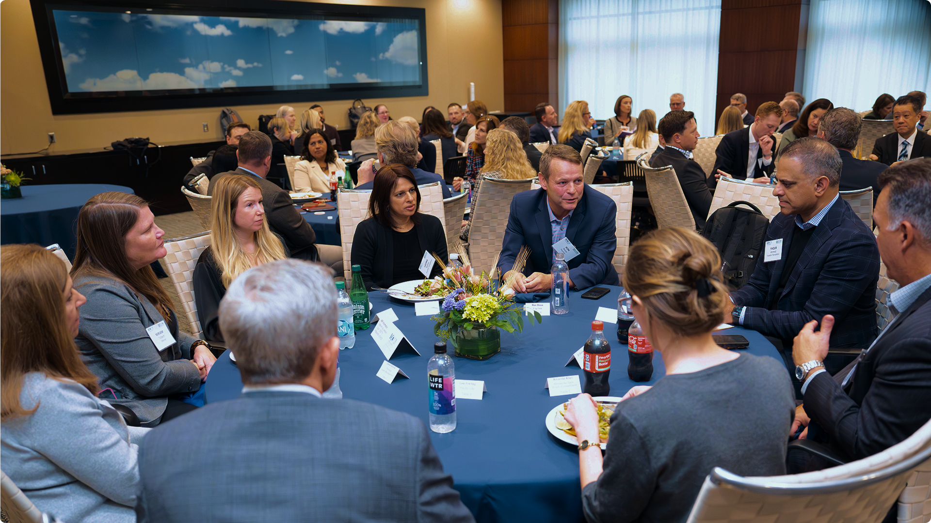 A group of business professionals engaged in conversation around a round table during a conference lunch.