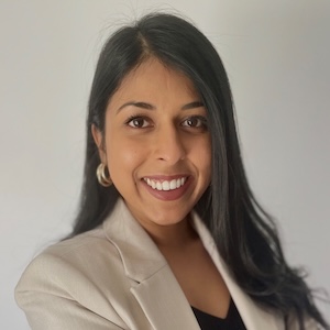 Smiling woman with long dark hair wearing a beige blazer and gold hoop earrings against a plain background.