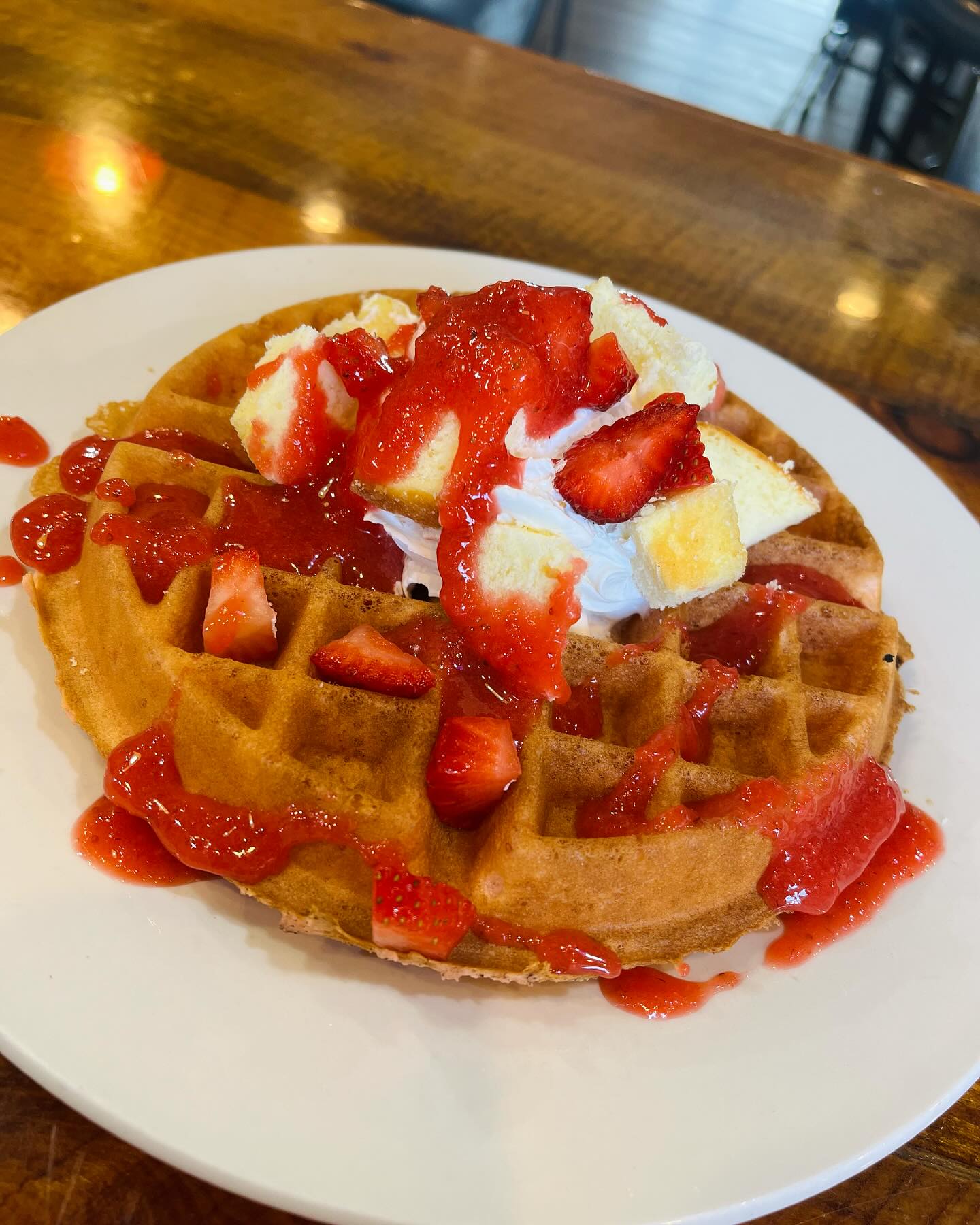 Plate with a golden waffle topped with whipped cream, cheesecake cubes, fresh strawberry pieces, and strawberry sauce drizzled over.
