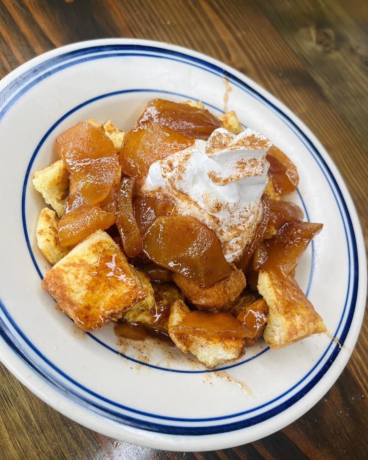 Plate of bread pudding topped with caramelized apple slices and whipped cream dusted with cinnamon on a wooden table.